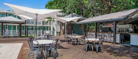 A wooden deck with outdoor seating and large umbrellas, surrounded by trees and a building with a bar area.