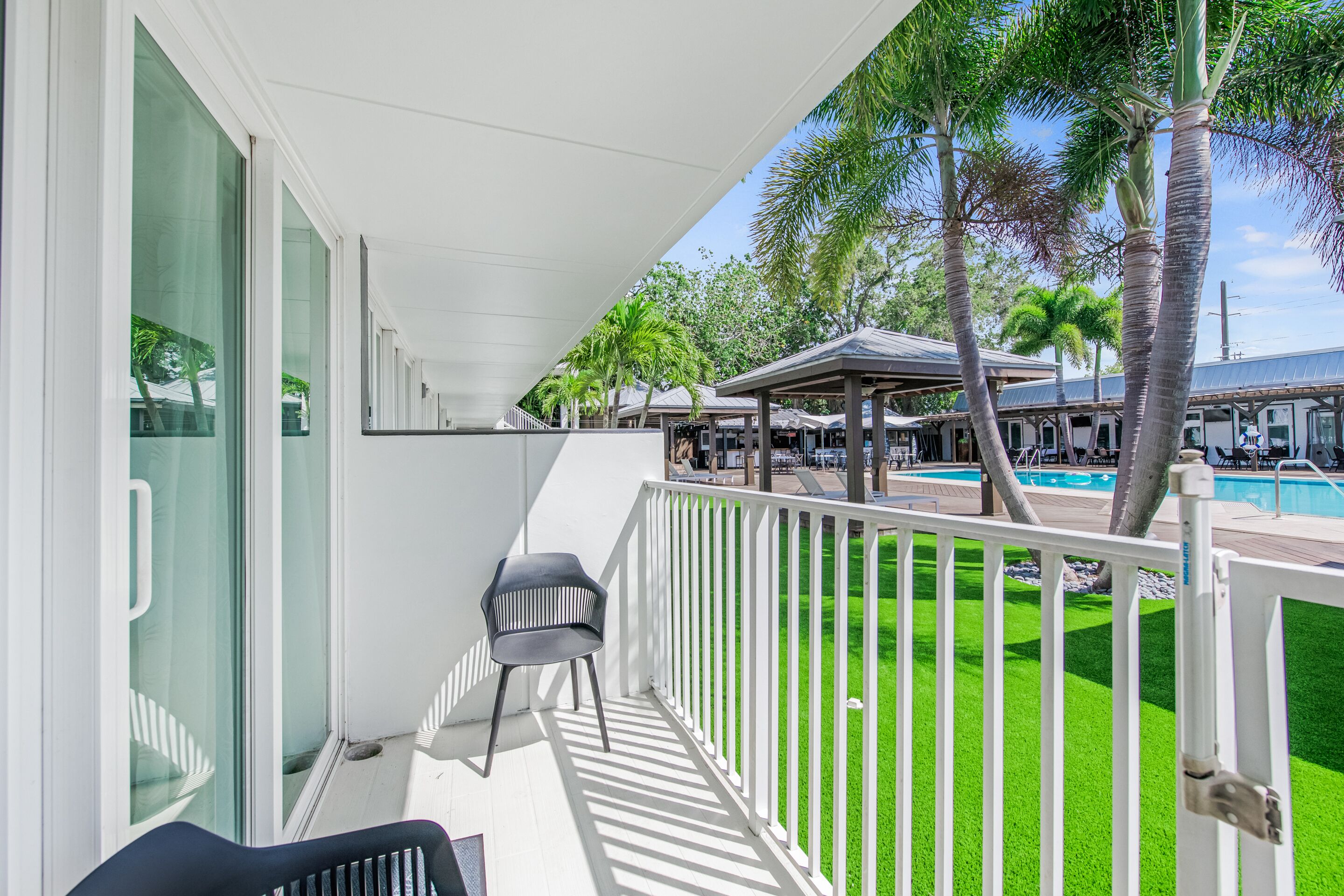 A balcony with a white railing, a black chair, and a view of a swimming pool and palm trees.