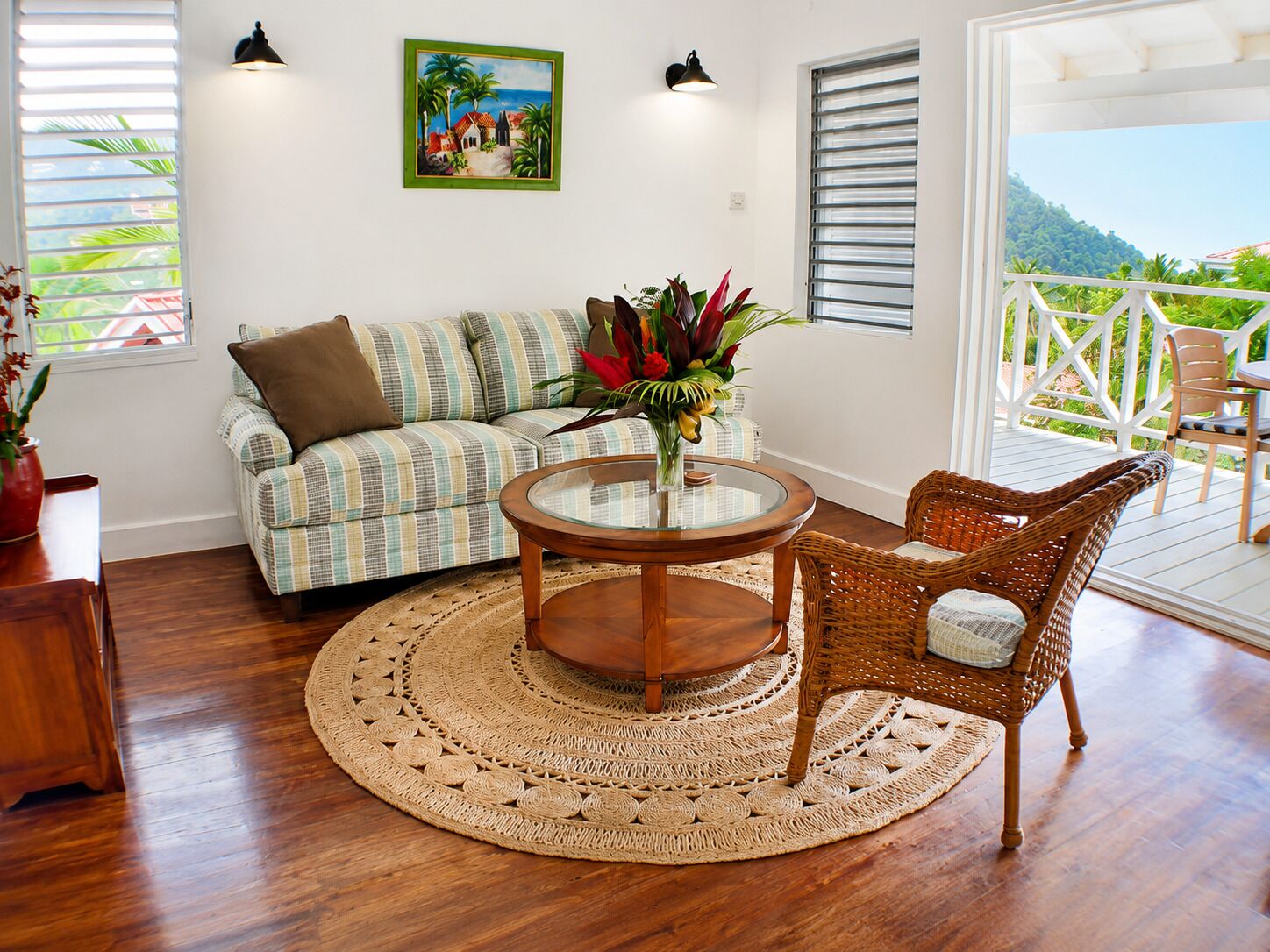 Quarto com sofá, mesa redonda, cadeira de vime e vista para uma varanda com mobiliário de exterior.