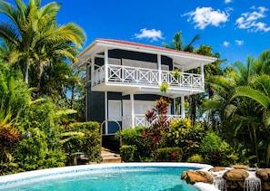 Un bâtiment à deux étages doté d’un balcon, entouré d’une végétation tropicale luxuriante et agrémenté d’une piscine.