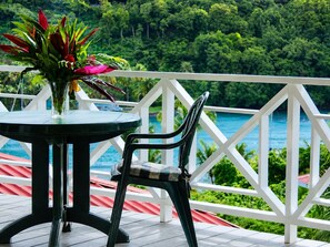 Un balcon avec une table et une chaise, donnant sur un paysage verdoyant et luxuriant et sur une étendue d’eau.