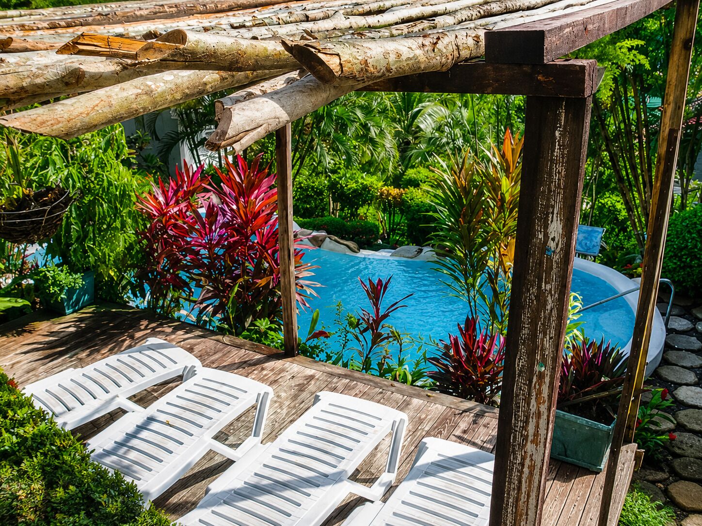 A wooden deck with white lounge chairs, a pool, and lush tropical plants.