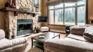 Una sala de estar con chimenea de piedra, un ventanal con vistas a árboles y un rincón acogedor con una mesita de centro.