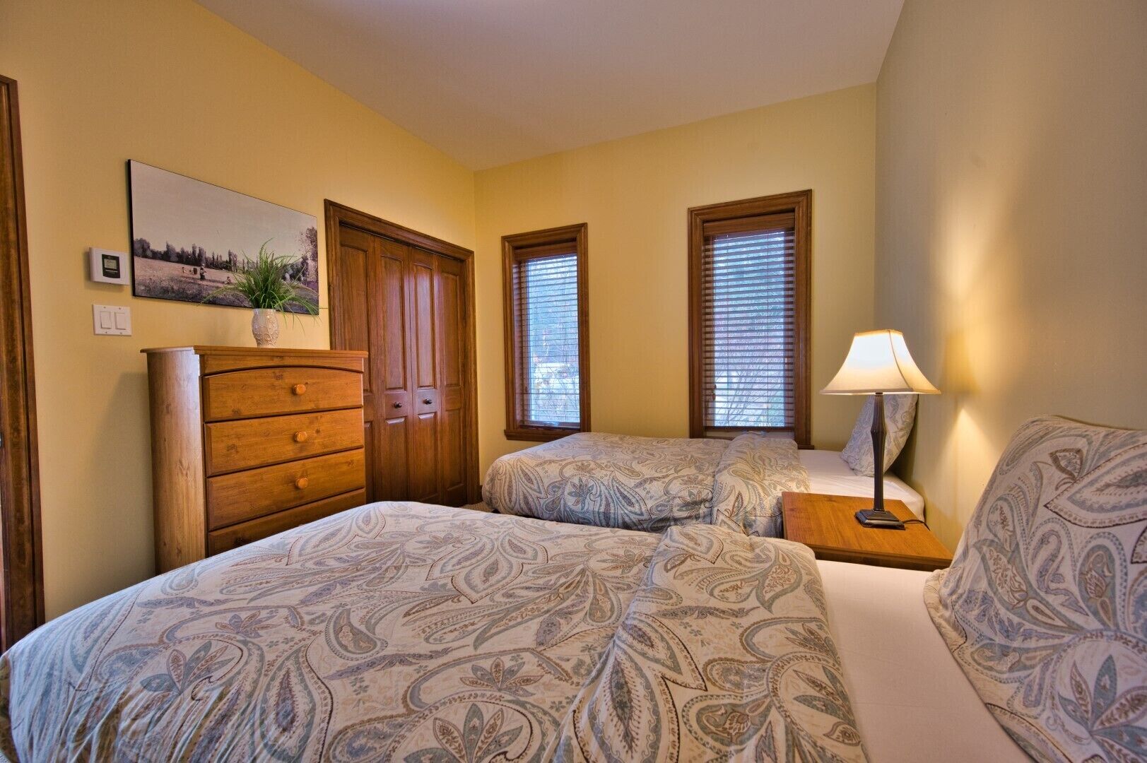 Une chambre avec deux lits, une commode en bois, une table de chevet avec une lampe et un tableau accroché au mur.