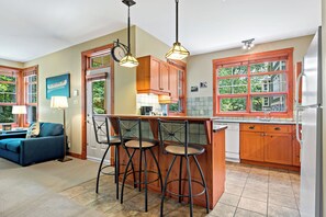 A kitchen with wooden cabinets, a bar area with stools, a clock, and a view of trees through large windows. - Tremblant-Les-Eaux – RVMT (Mont-Tremblant)