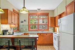 A kitchen with wooden cabinets, a large window, a refrigerator, and a bar area with stools. - Tremblant-Les-Eaux – RVMT (Mont-Tremblant)