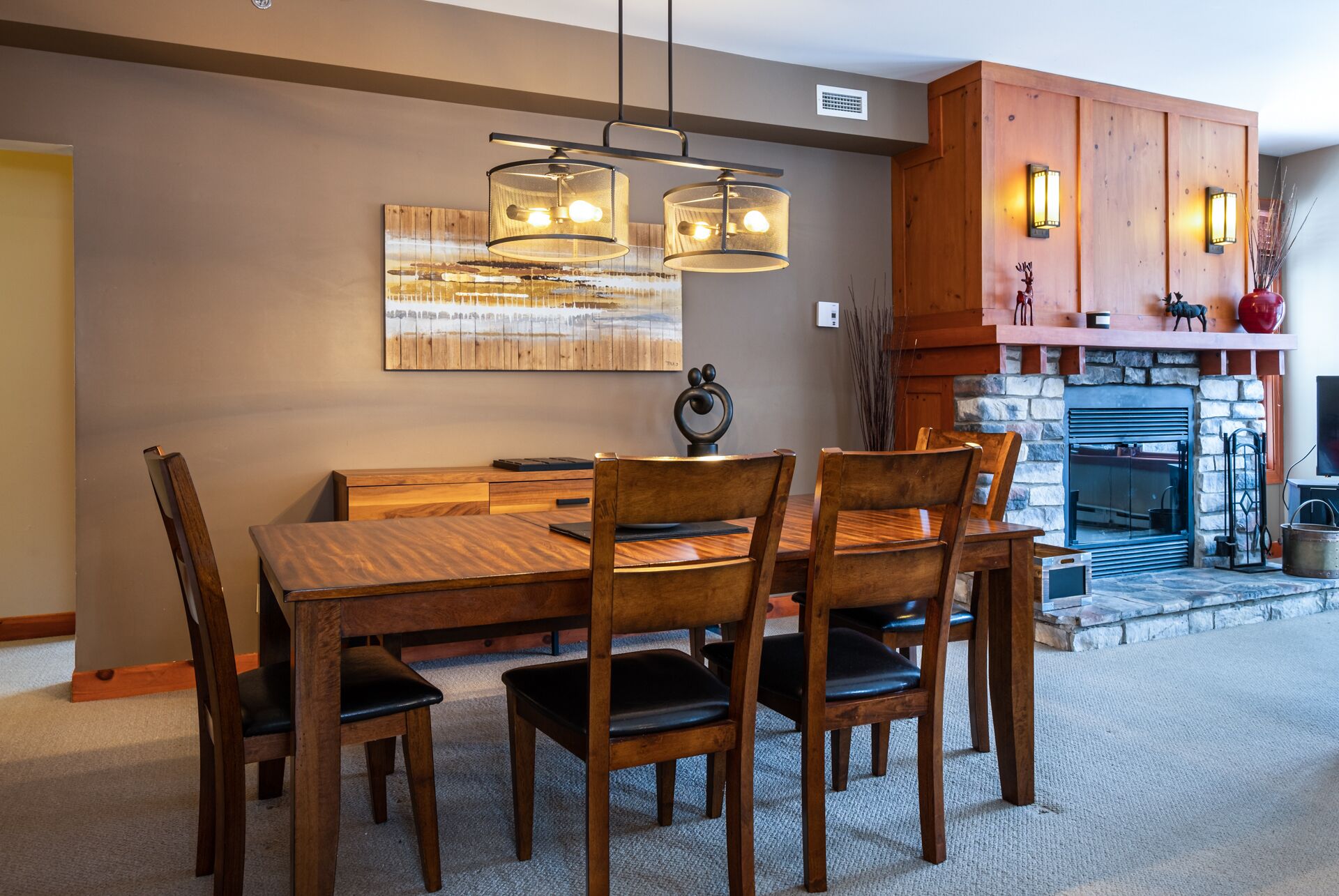 A dining area with a wooden table and chairs, a hanging light fixture, and a fireplace with stone walls.