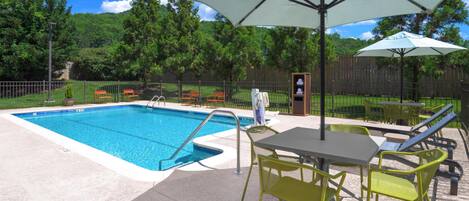 A swimming pool with a sunshade, outdoor seating, and a fenced area with trees in the background.
