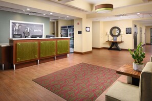 A hotel reception area with a reception desk, a refrigerator, a mirror, and a potted plant.