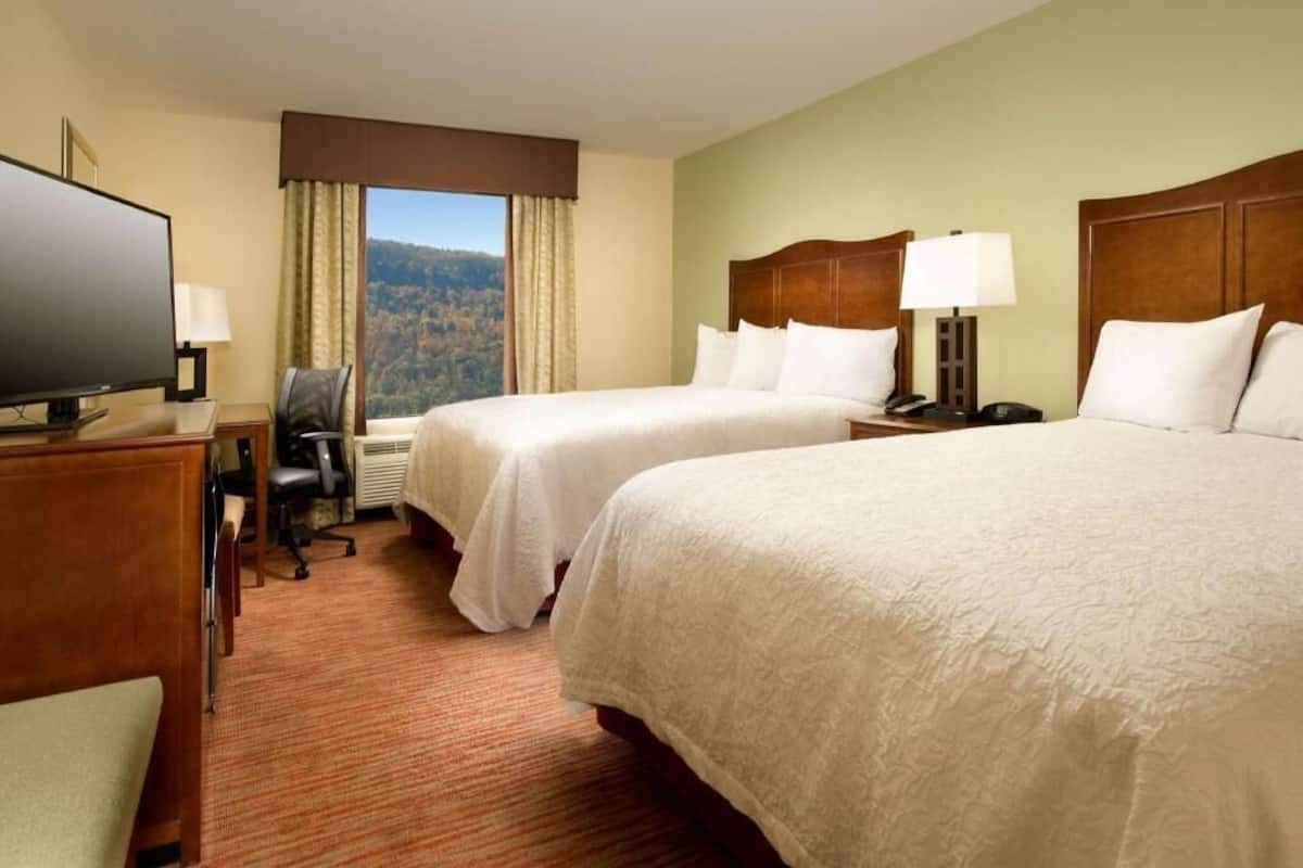 A hotel room with two beds, a desk with a television, a chair, and a window with a mountain view.