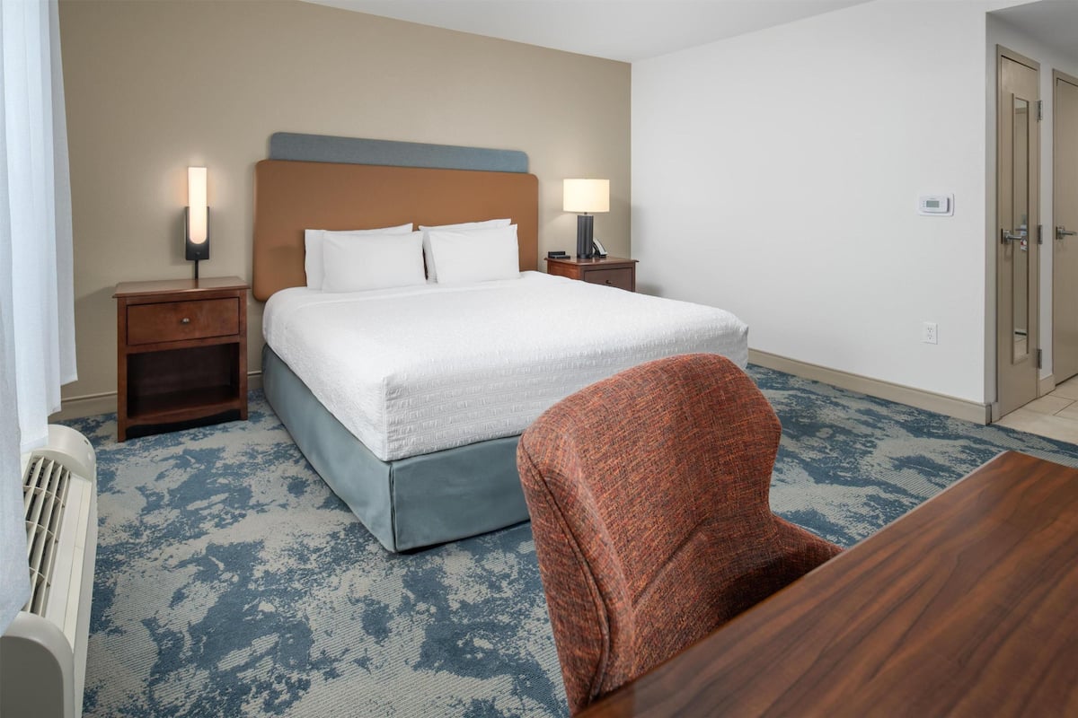 A hotel room with a large bed, two bedside tables with lamps, a wooden nightstand, a red chair, and a wooden table.