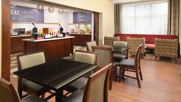 A hotel dining area with a central table, multiple chairs, a kitchenette, and a seating area with patterned chairs.