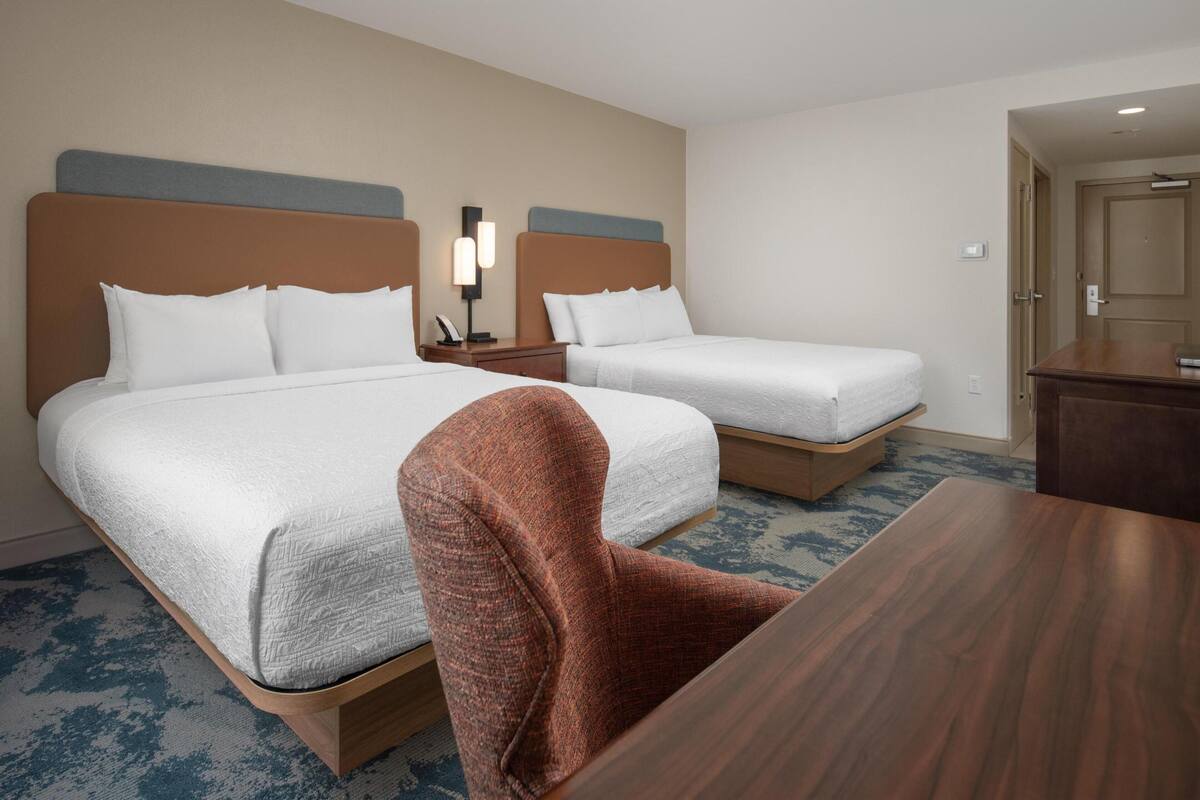 A hotel room with two beds, a wooden nightstand, a table, and a chair.