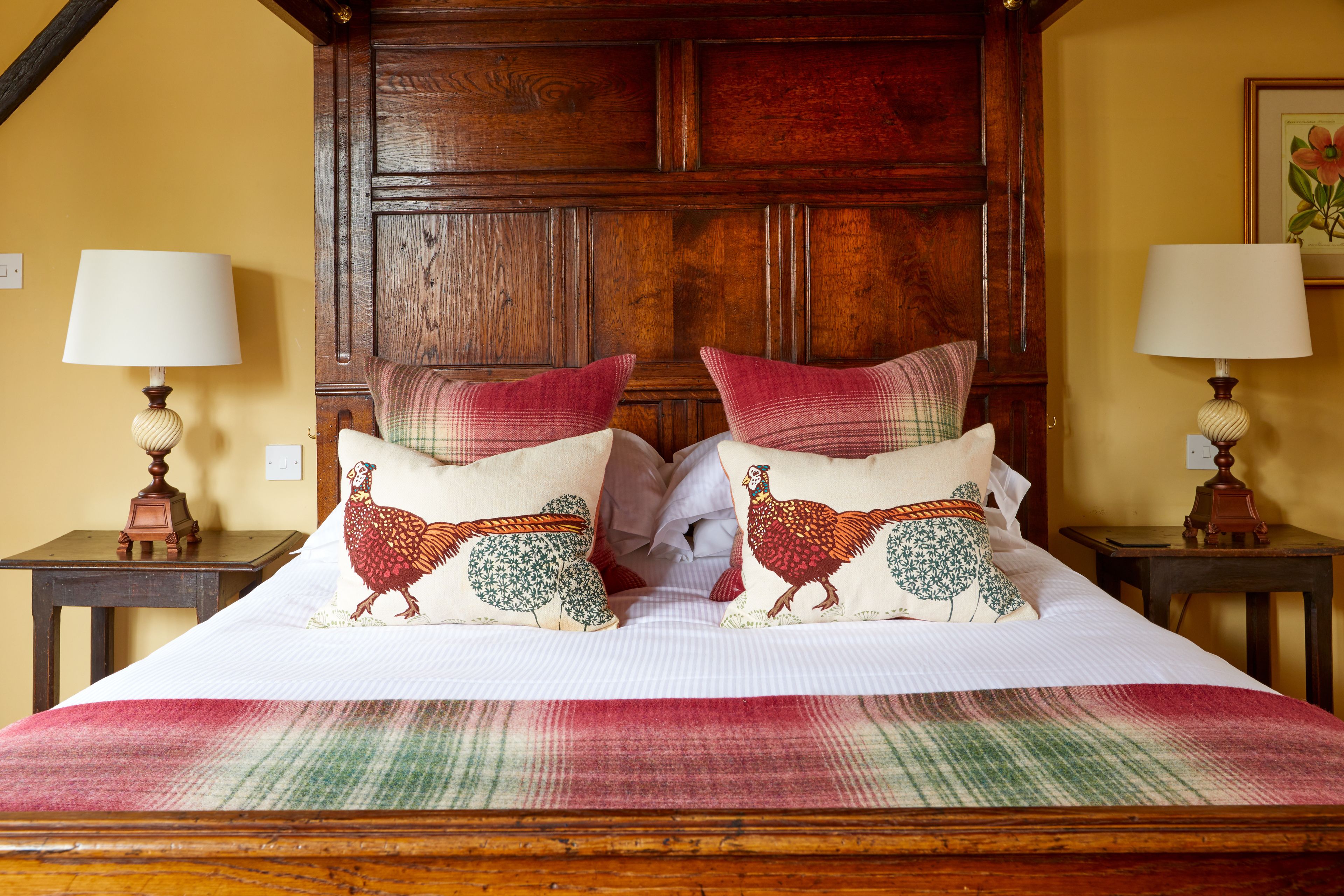 Una cama con cabecera de madera, dos mesitas de noche con lámparas y almohadones decorados con pavo.
