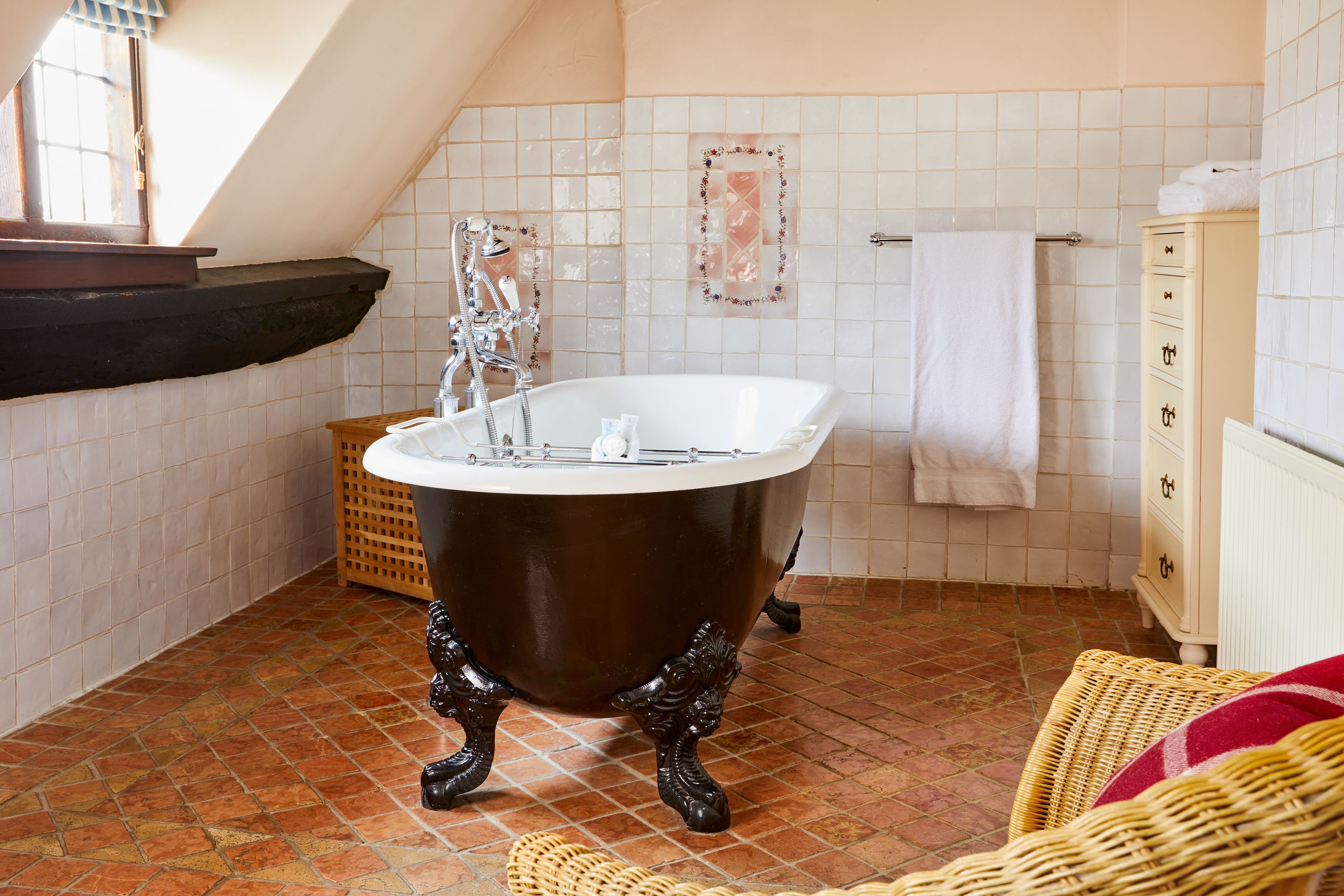 A bathroom with a clawfoot bathtub, a wooden basket, a wicker chair, and a tiled floor.