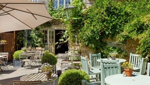 An outdoor patio area with multiple tables and chairs, surrounded by greenery and a stone wall. - Burford House (Burford)