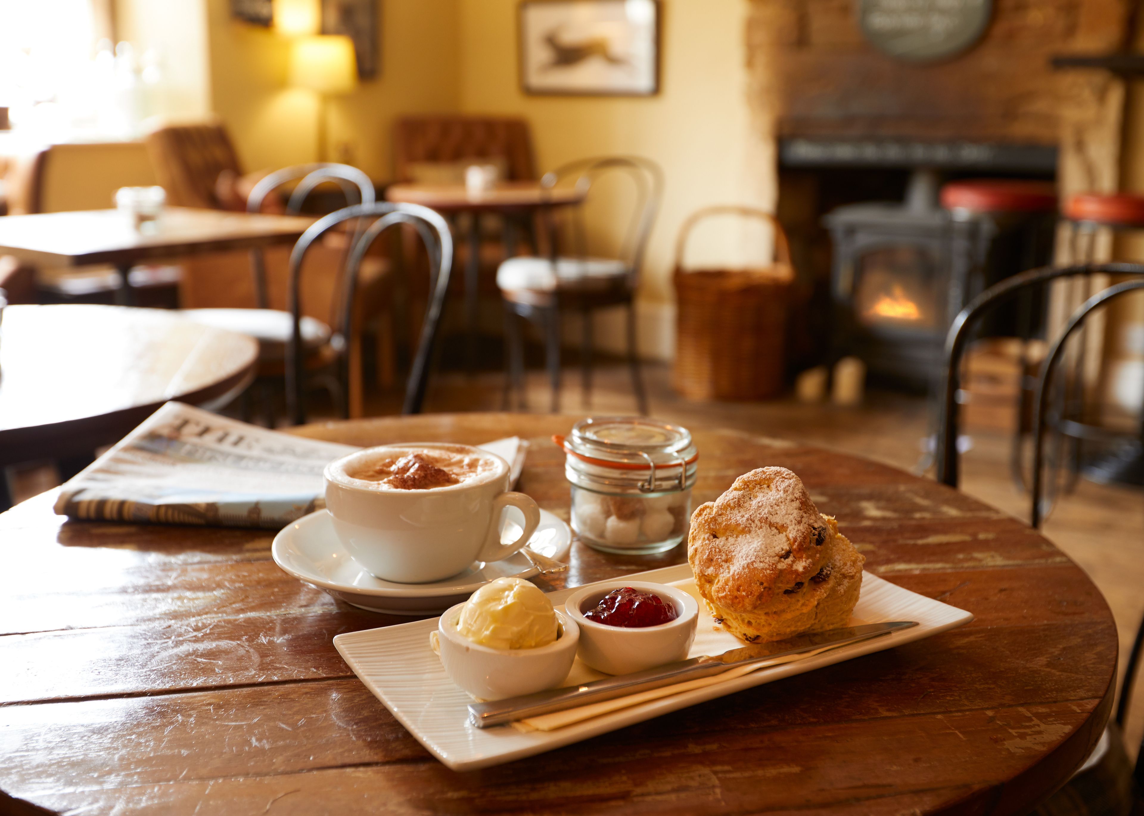 A cozy dining area with a round wooden table, a cup of coffee, a pastry, and a small bowl of jam.