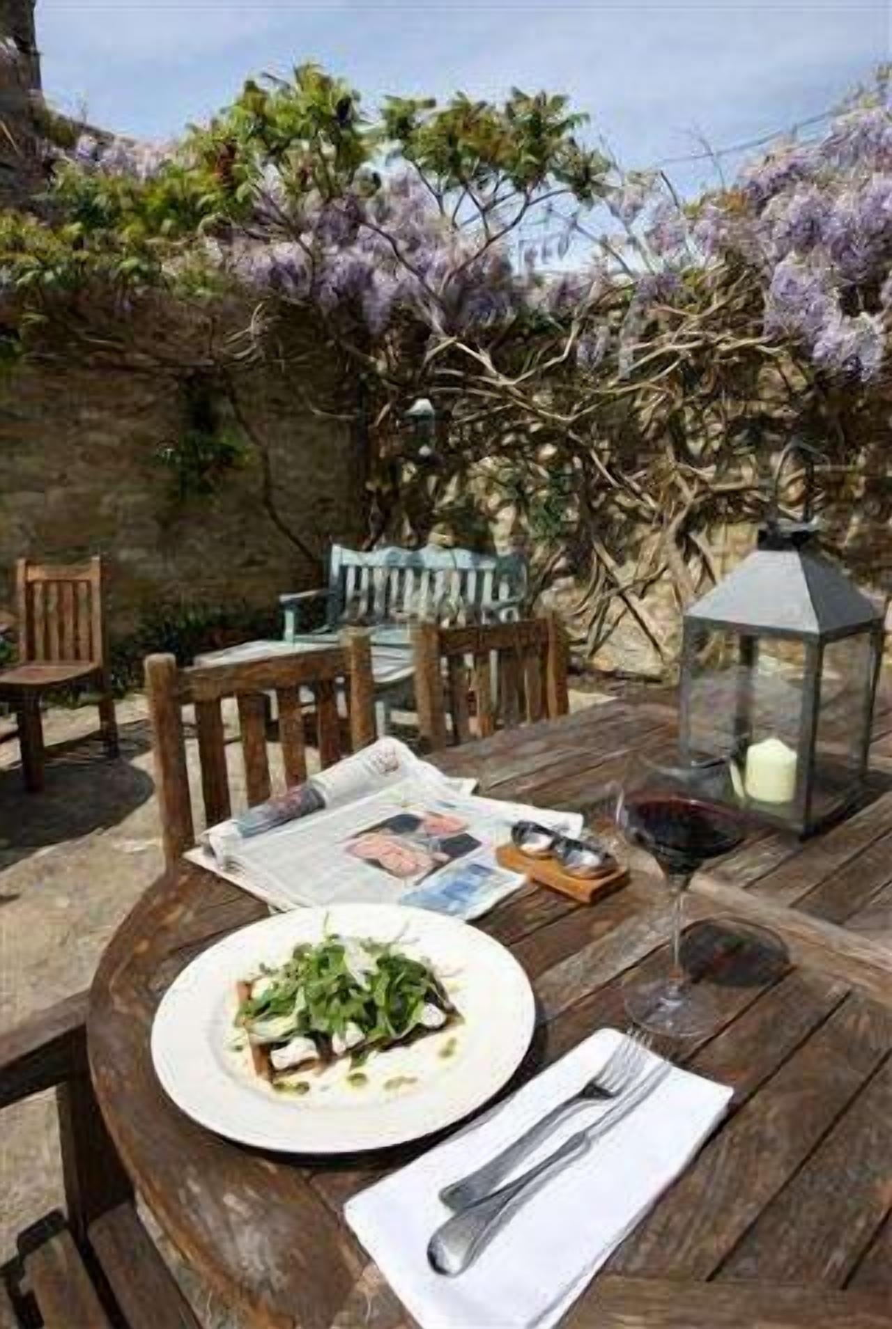 A wooden table set with a plate of food, a wine glass, and a candle in a garden with a stone wall and flowering trees.