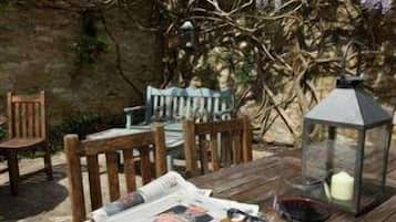 A wooden table set with a plate of food, a wine glass, and a candle in a garden with a stone wall and flowering trees.
