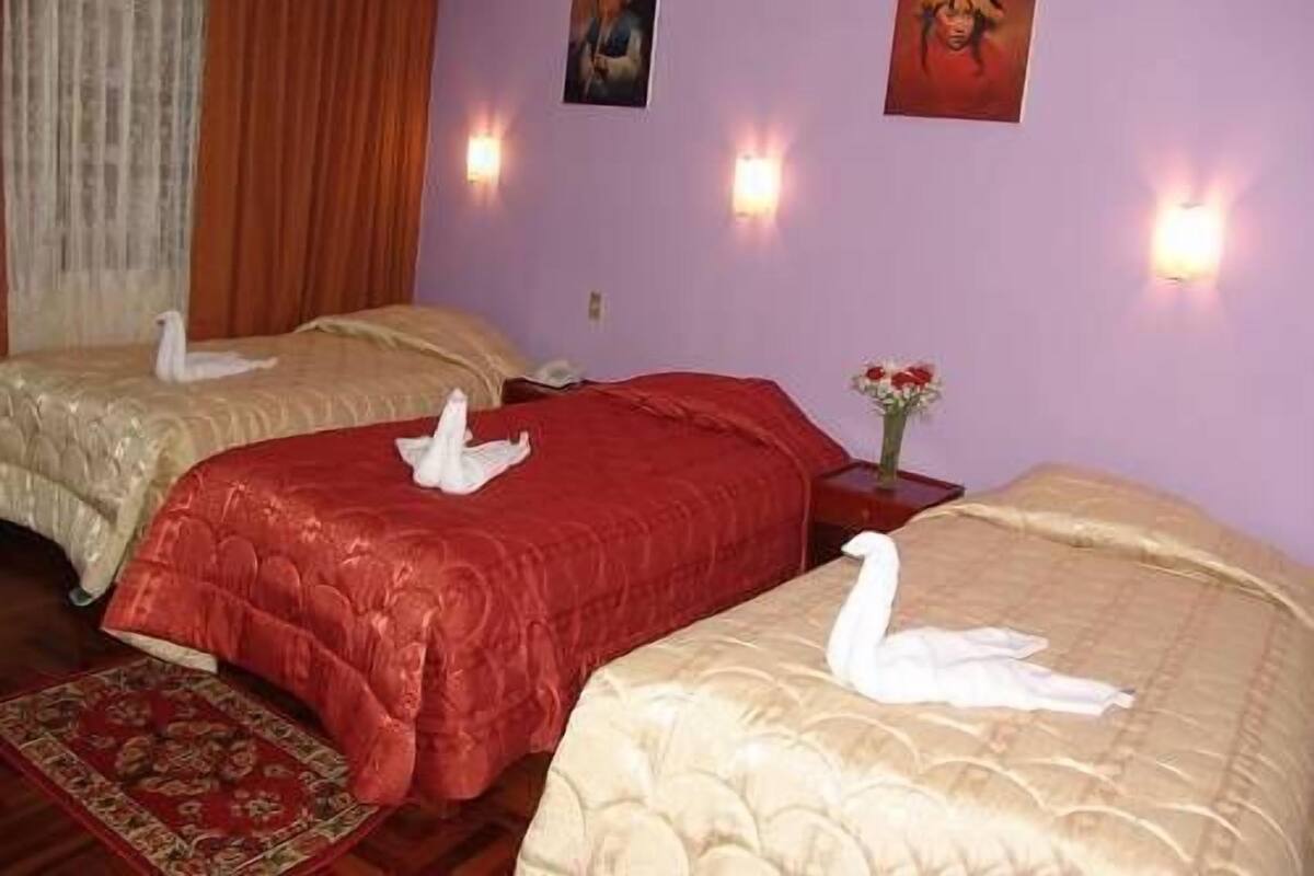 A hotel room with two beds, each with a swan-shaped pillow, a red bedspread, and a beige bedspread. There are wall-mounted lights and a vase with flowers on a nightstand.