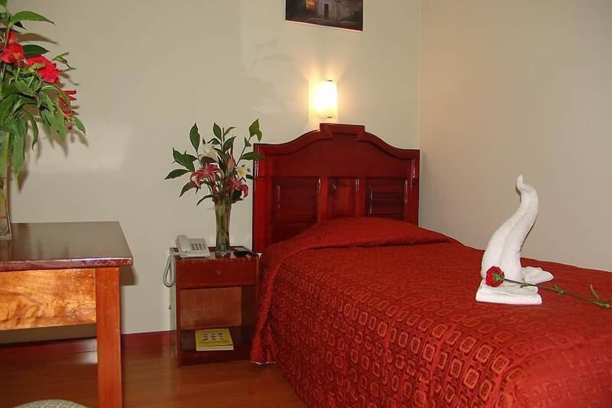 A hotel room with a wooden bed, a bedside table with a lamp, a vase with flowers, and a painting on the wall.