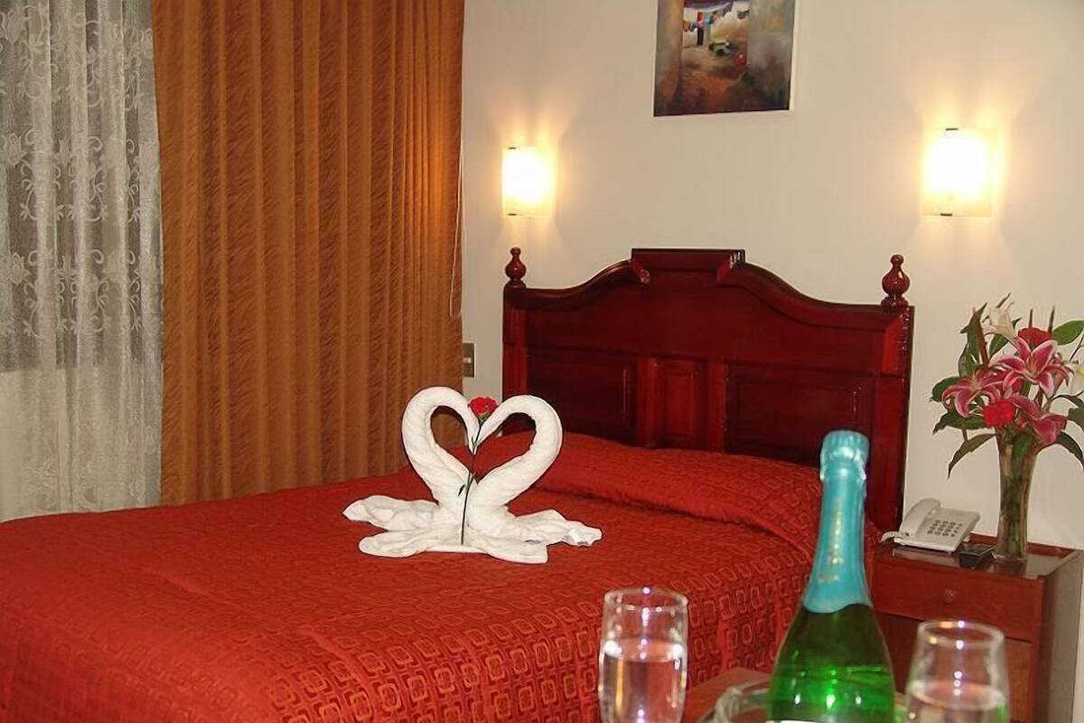 A hotel room with a red bedspread, a wooden headboard, two wall sconces, a bouquet of flowers, a bottle of champagne, and two glasses on the nightstand.