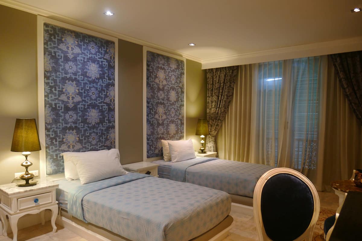Habitación de hotel con dos camas, una mesita de noche con lámpara, una silla y vistas al exterior por la ventana.
