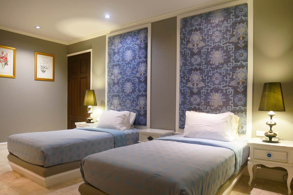 Una habitación de hotel con dos camas, una mesita de noche con lámpara y una puerta que da a otra habitación.