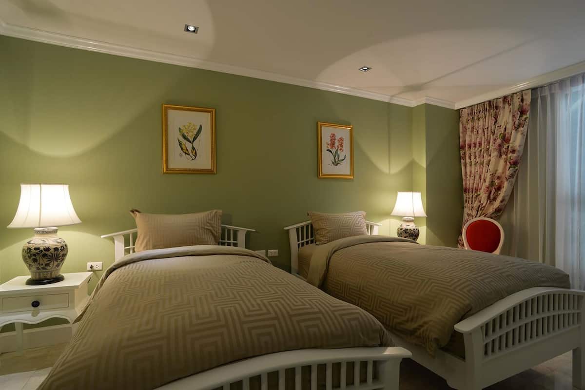 Una habitación con dos camas, paredes verdes, impresiones botánicas enmarcadas, lámparas de noche y un cortinaje floral.