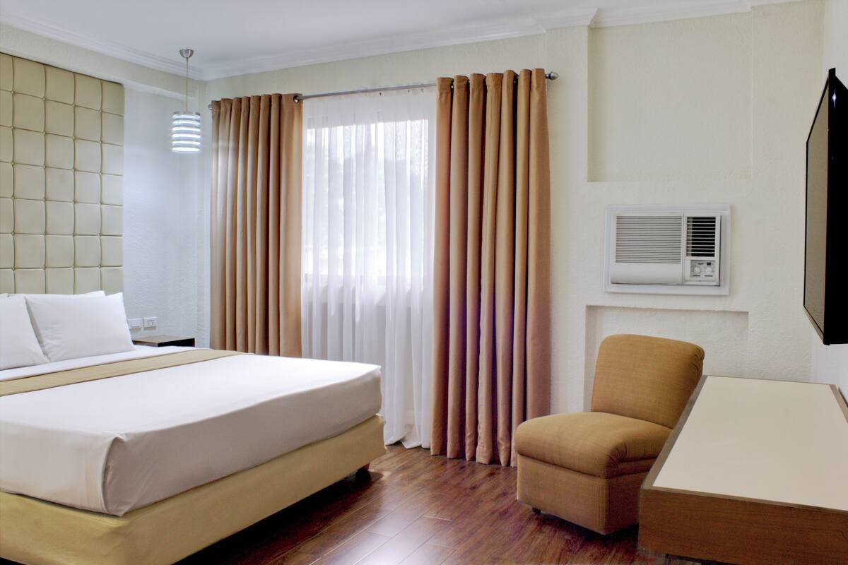 A hotel room with a bed, a chair, a desk, curtains, and an air conditioning unit.