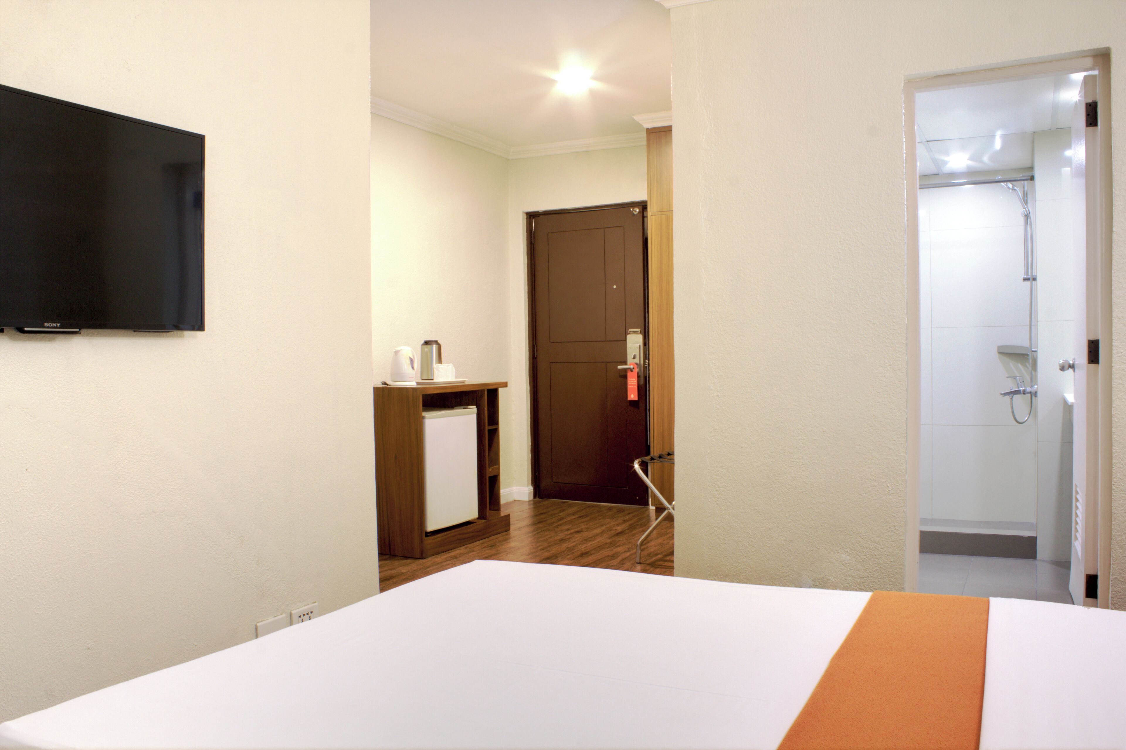 Habitación de hotel con una cama, un pequeño escritorio con una tetera, un televisor montado en la pared y una zona de ducha visible a través de una puerta abierta.