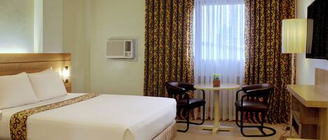 Quarto de hotel com uma cama grande, uma mesa pequena com duas cadeiras, ar condicionado e uma televisão.