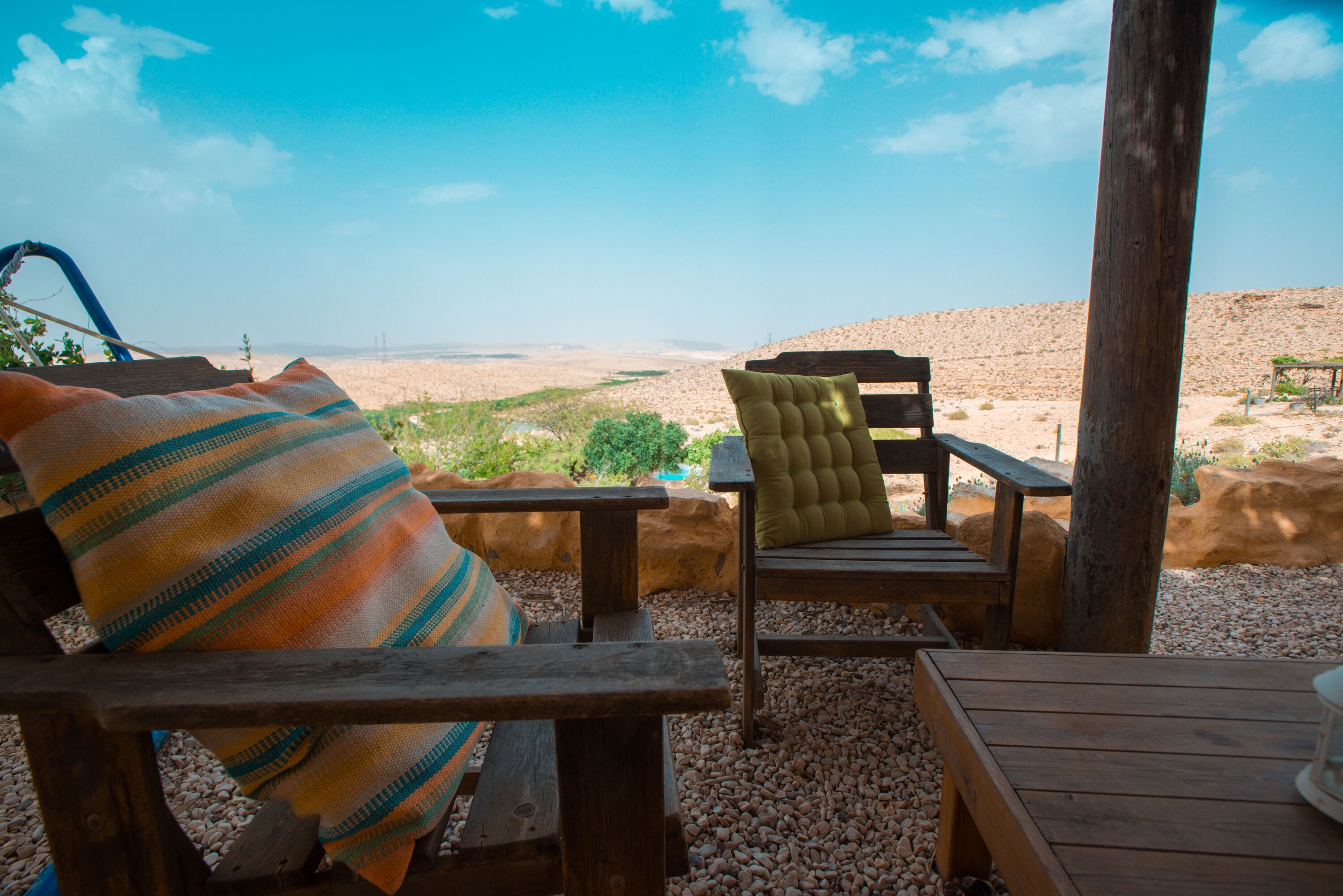 Eine Holzterrasse mit zwei Stühlen, einer mit einer Streifen-Decke und der andere mit einem grünen Kissen, mit Blick auf eine Wüstenlandschaft.