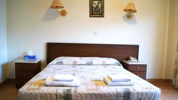 Cama bem arrumada com colcha, duas mesas de cabeceira com abajures, um quadro na parede e um móvel pequeno com gavetas.