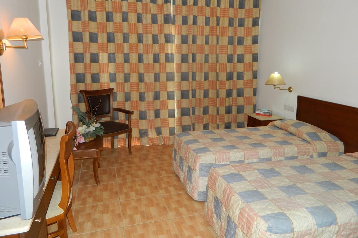 A hotel room with a checkered curtain, a bed with a quilt, a chair, a small table with a lamp, and a television.