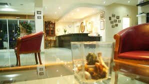 A lobby with a reception desk, two red leather chairs, a glass display case with items, and a tiled floor. - Moniatis Hotel (Limassol)