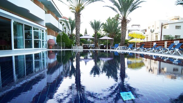 A swimming pool with lounge chairs and umbrellas, surrounded by palm trees and buildings. - Moniatis Hotel (Limassol)
