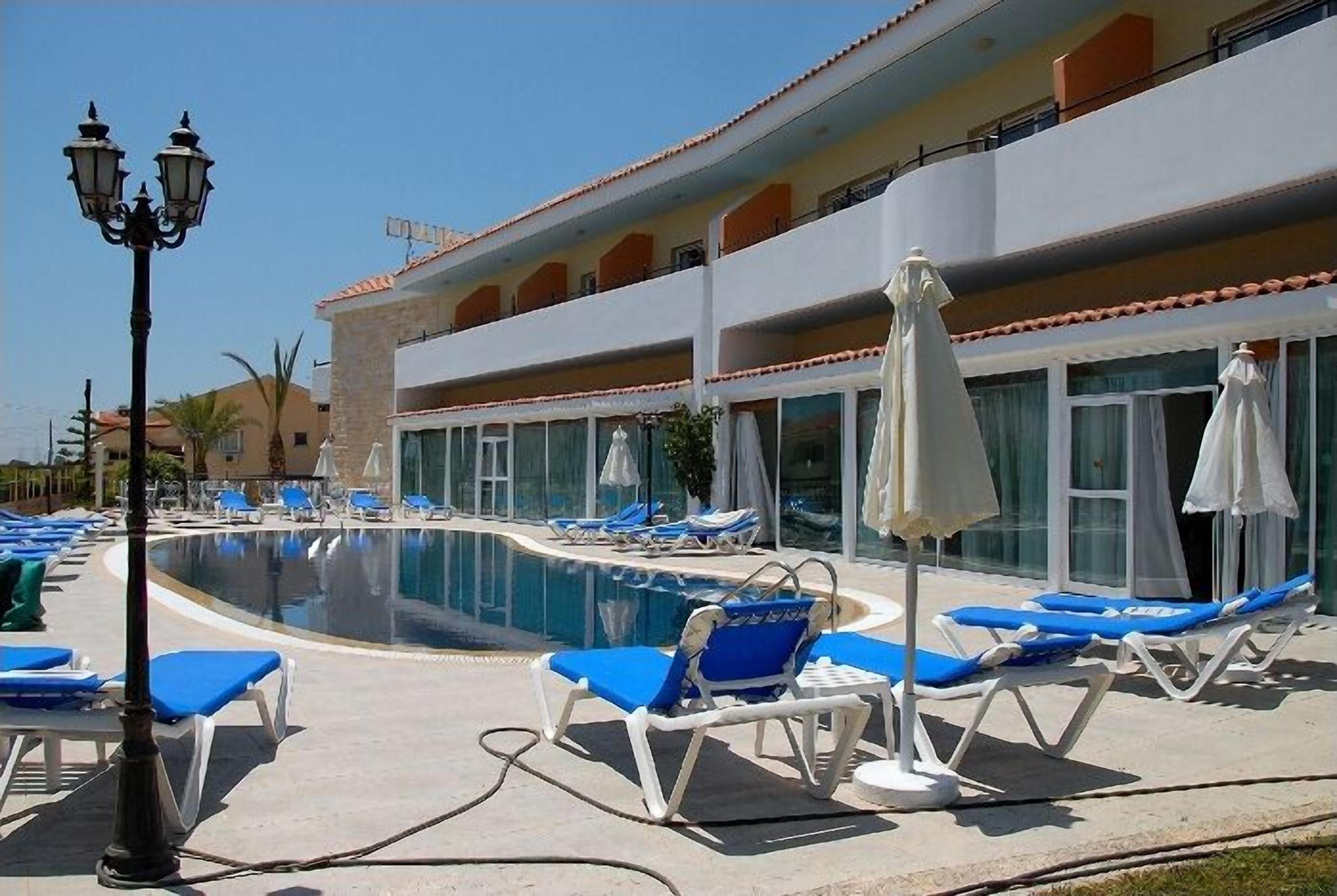 Zona junto a la piscina con sillones de descanso y sombrillas, un edificio con balcones y un cielo despejado.