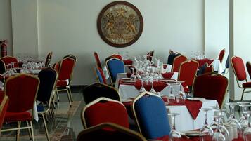 Un salón de banquetes con mesas dispuestas para un evento formal, con sillas rojas y azules, manteles blancos y un escudo en la pared.