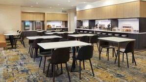 A hotel breakfast area with tables and chairs, a coffee machine, and a breakfast bar with various food items. - TownePlace Suites by Marriott Thunder Bay (Thunder Bay)