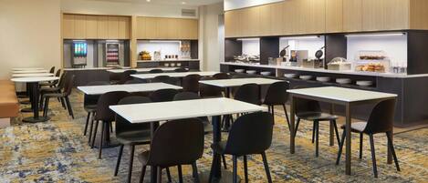 A hotel breakfast area with tables and chairs, a coffee machine, and a breakfast bar with various food items.