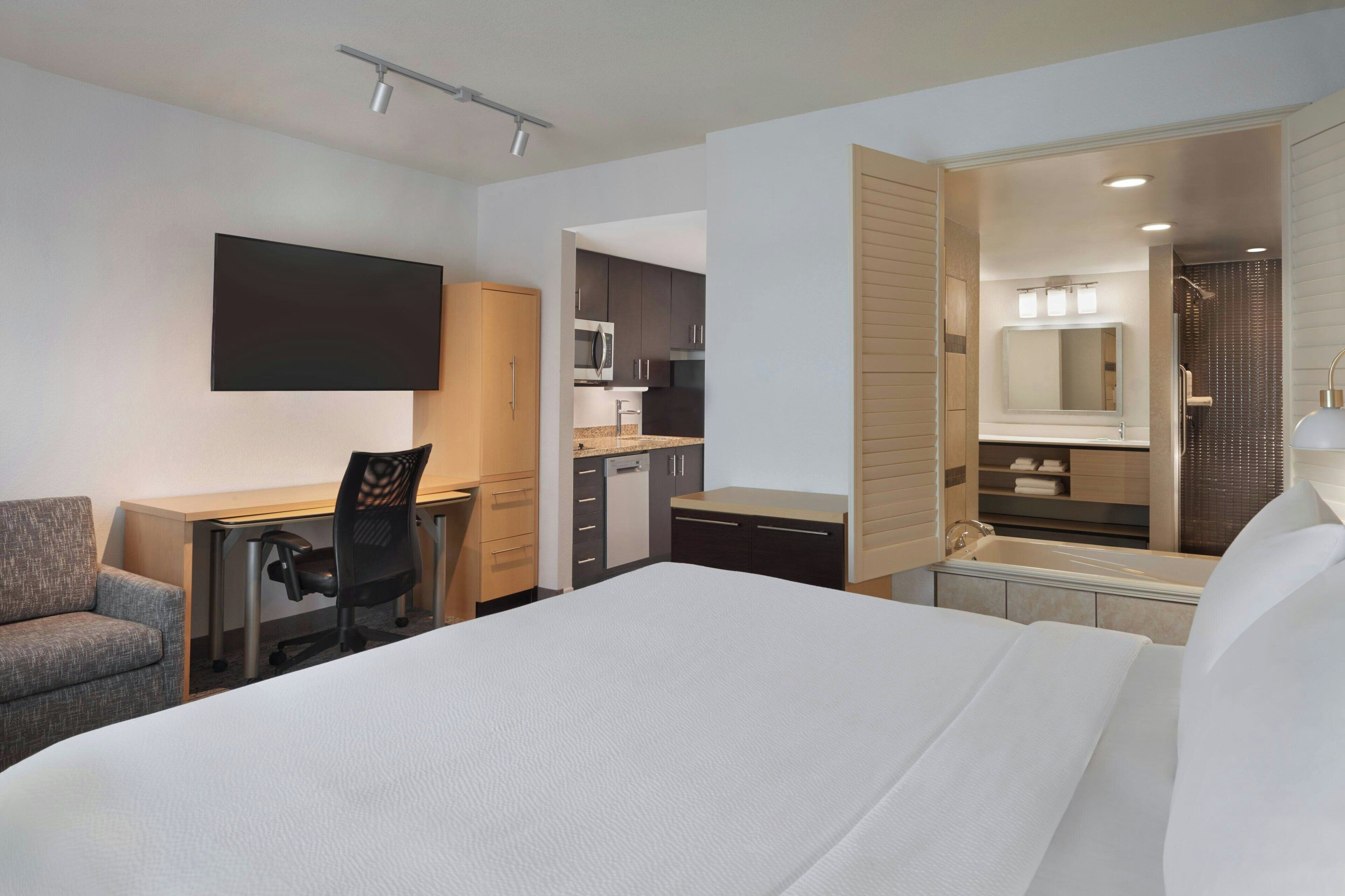 A hotel room with a bed, a desk, a chair, a kitchenette, and a bathroom.