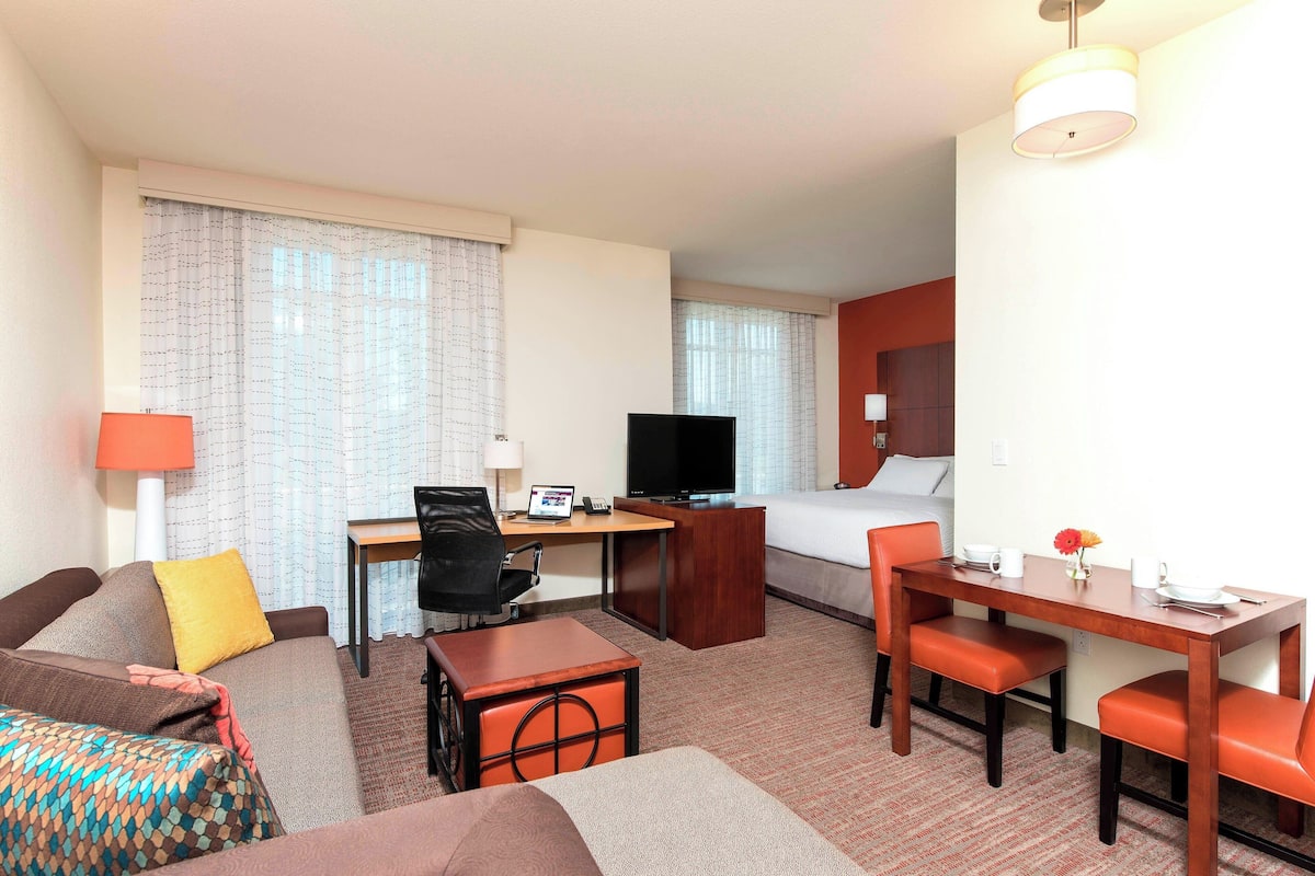 A hotel room with a bed, a desk with a computer, a TV, a sofa, and a dining table with chairs.