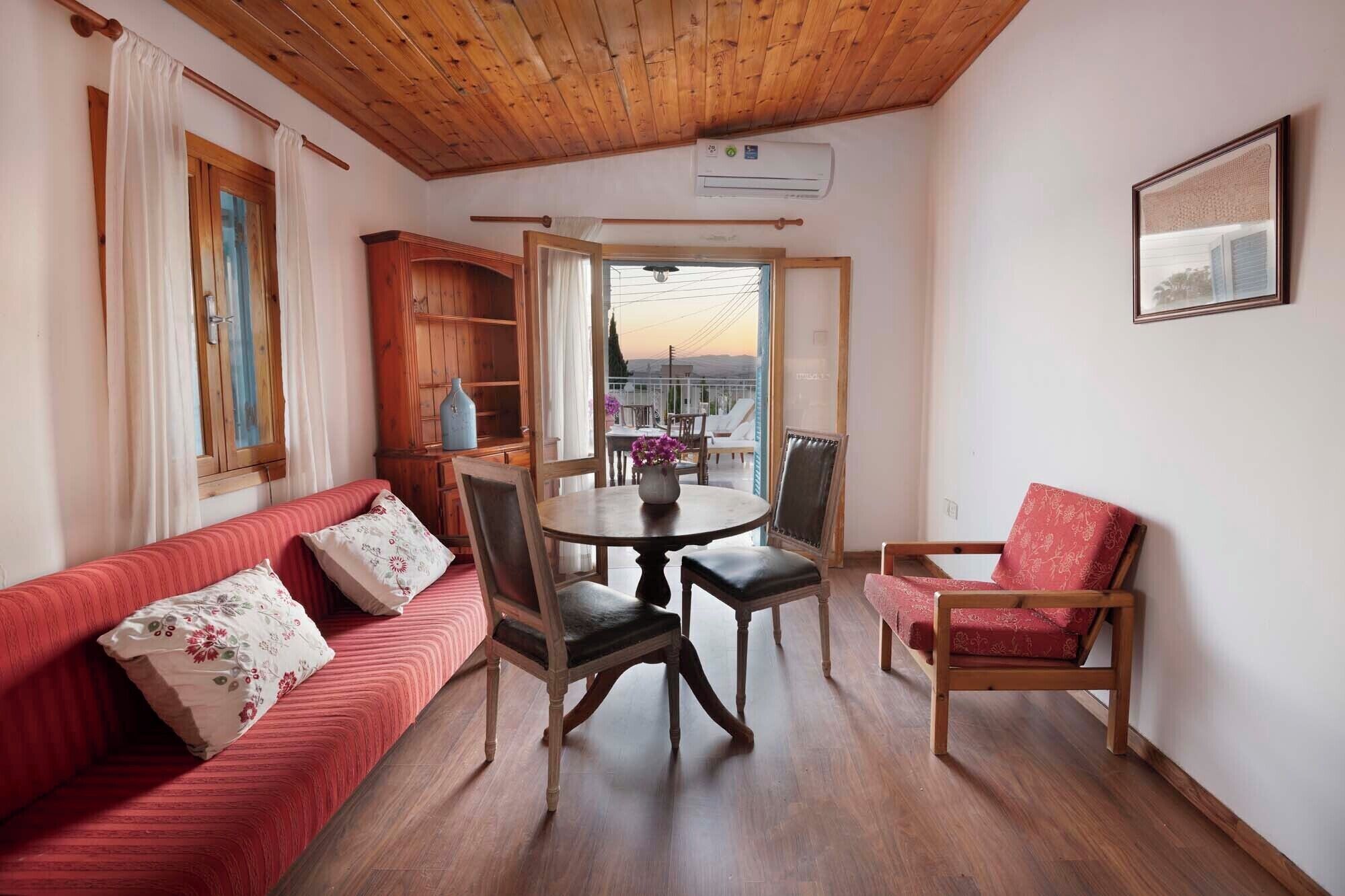 A cozy room with a wooden ceiling, a red sofa with floral cushions, a wooden dining table with chairs, and a view through a glass door to the outside.
