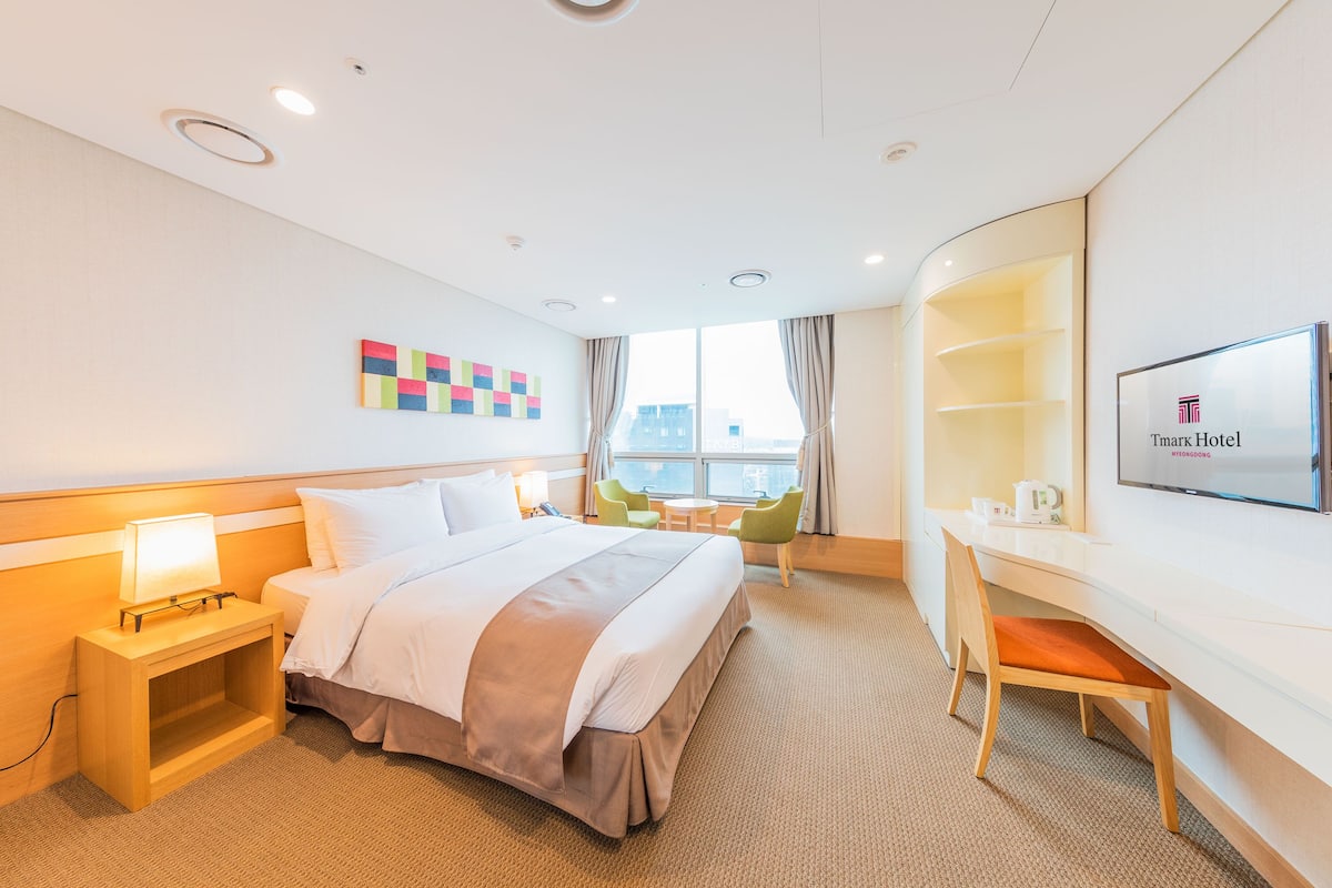 Habitación de hotel con cama, mesita de noche, lámpara, escritorio, silla, televisor y ventana con cortinas.