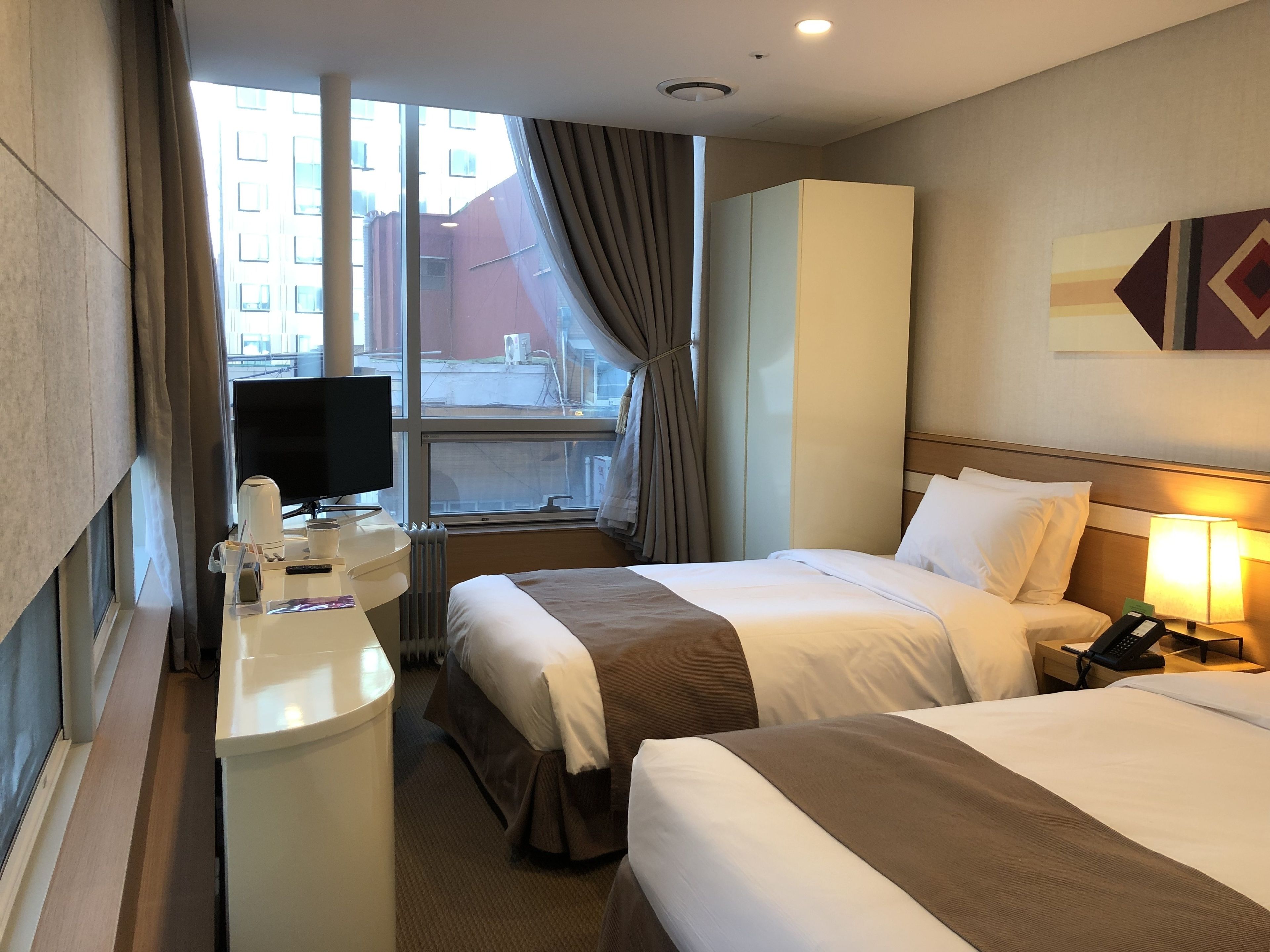 A hotel room with two beds, a desk with a computer and a coffee maker, a TV, and a window with curtains.