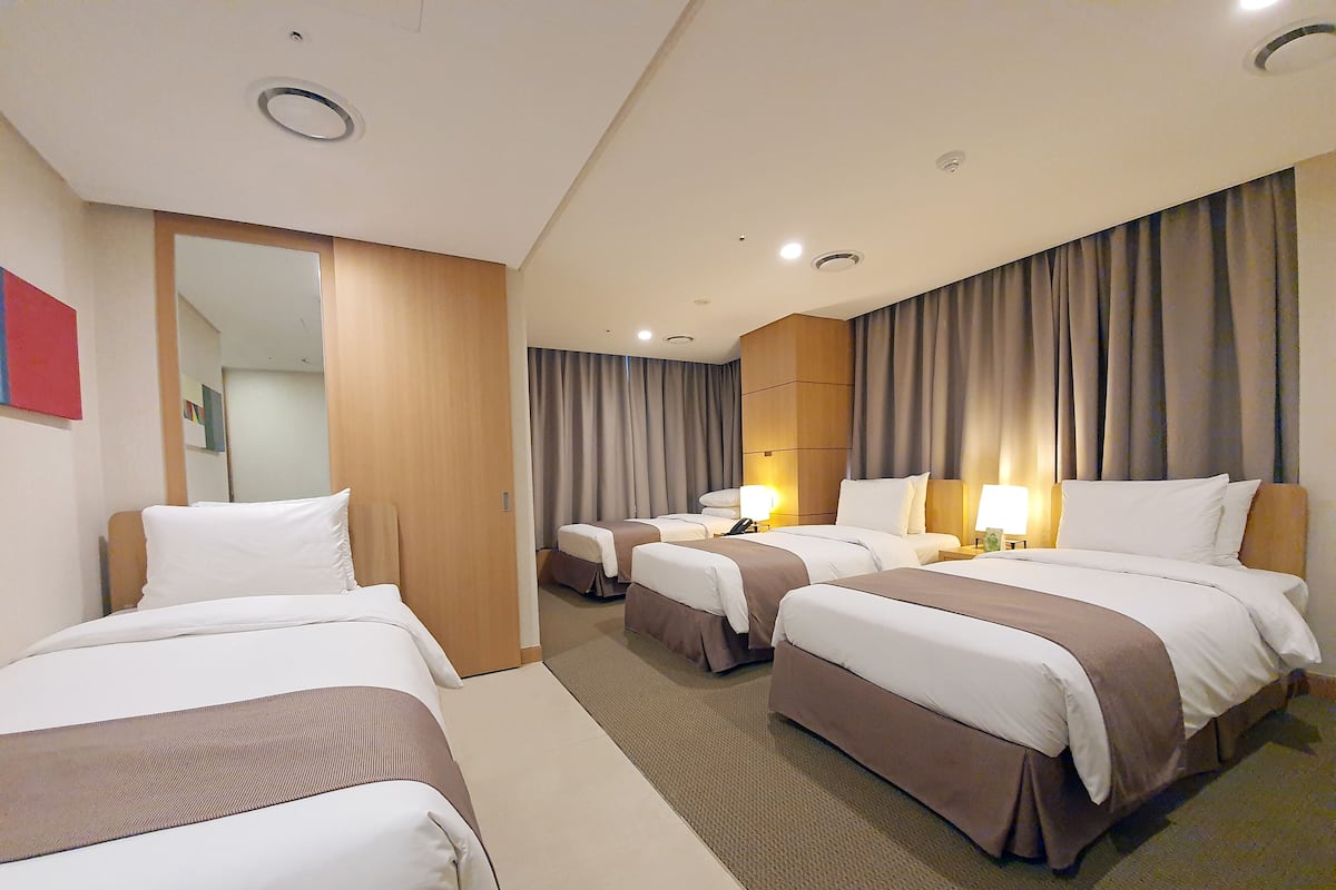 Habitación de hotel con cuatro camas dispuestas en fila, un espejo grande y vistas al exterior a través de la ventana.