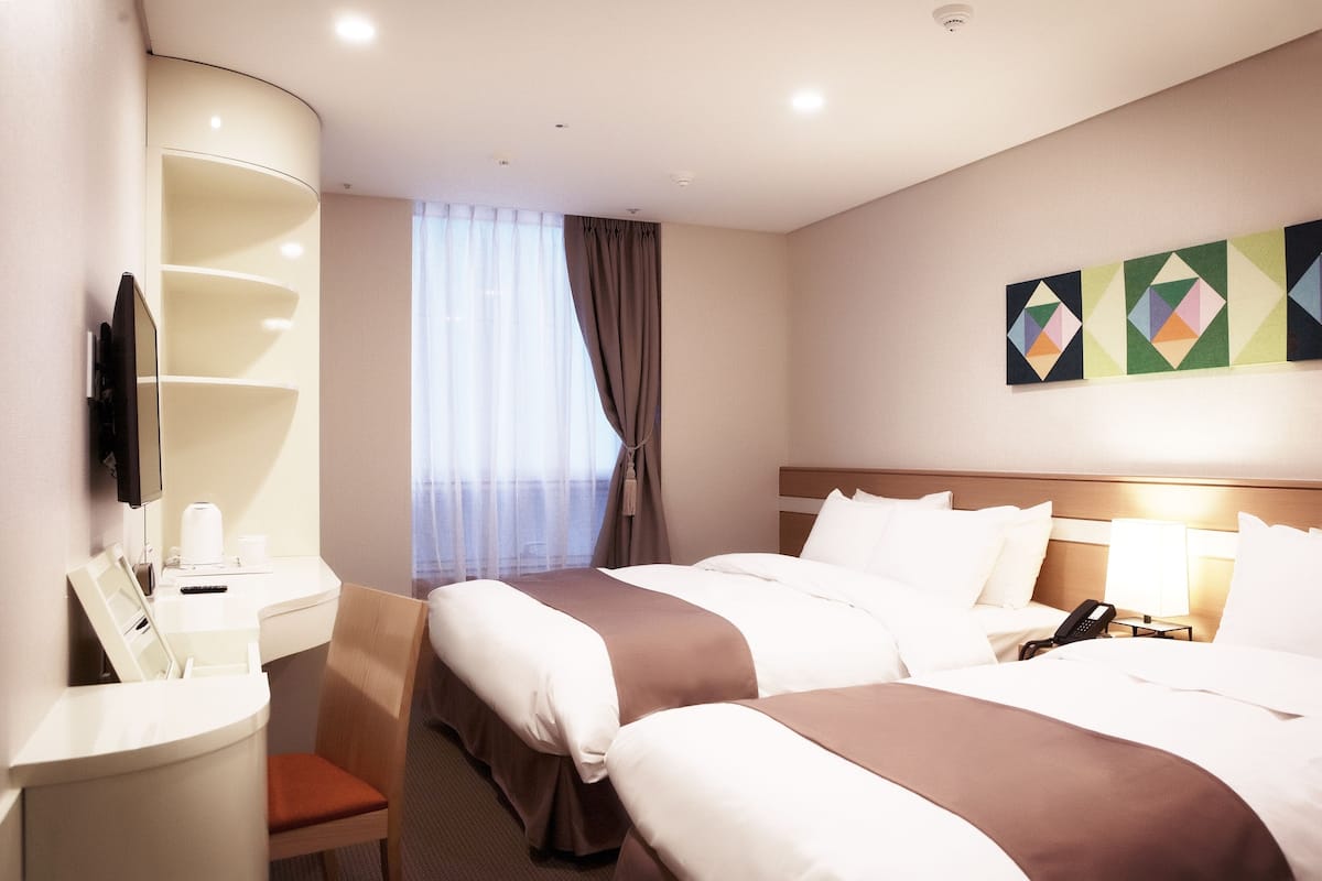Habitación de hotel con dos camas, un escritorio, una silla, un televisor y obras de arte en la pared.