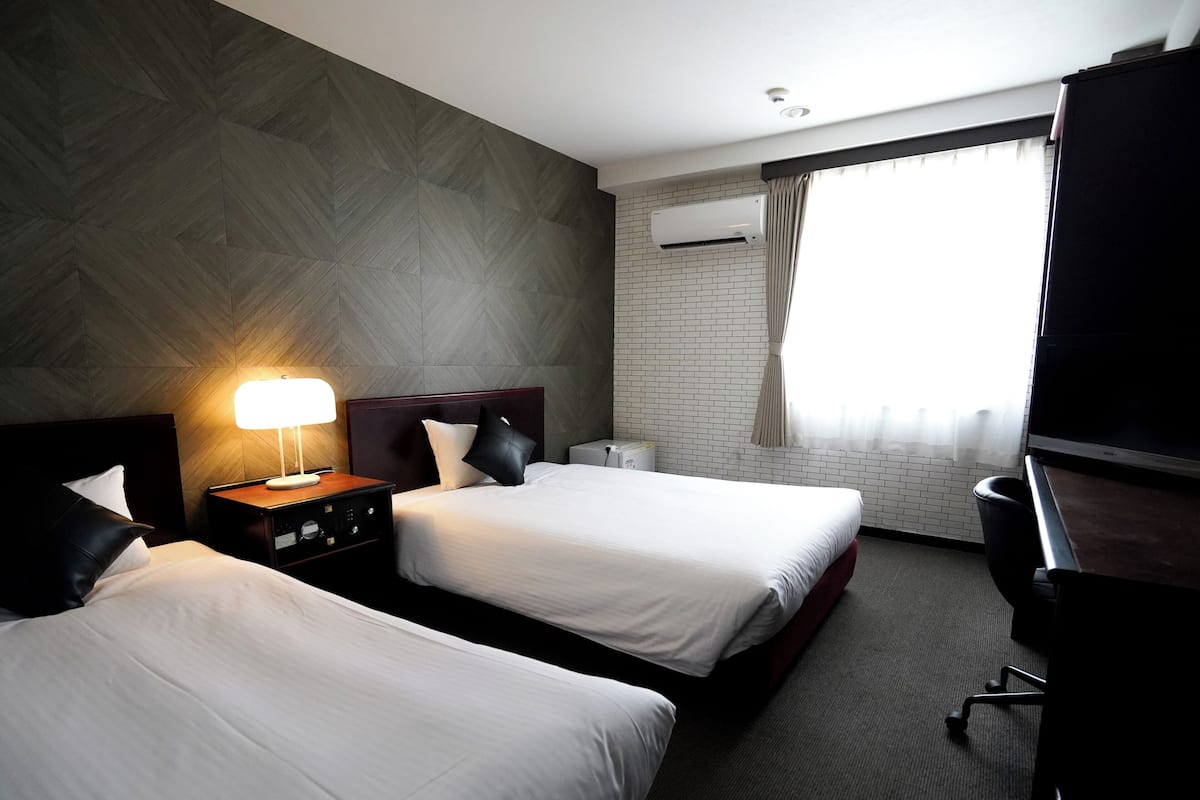 Habitación de hotel con dos camas, una mesita de noche con lámpara, un escritorio con silla, un equipo de aire acondicionado y una ventana con cortinas.