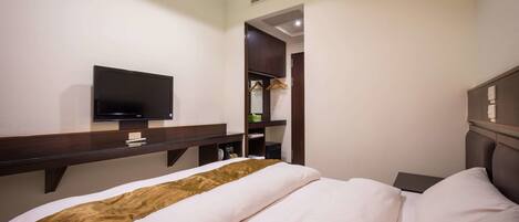 A hotel room with a bed, a TV mounted on a wooden shelf, a wardrobe, and a small table.